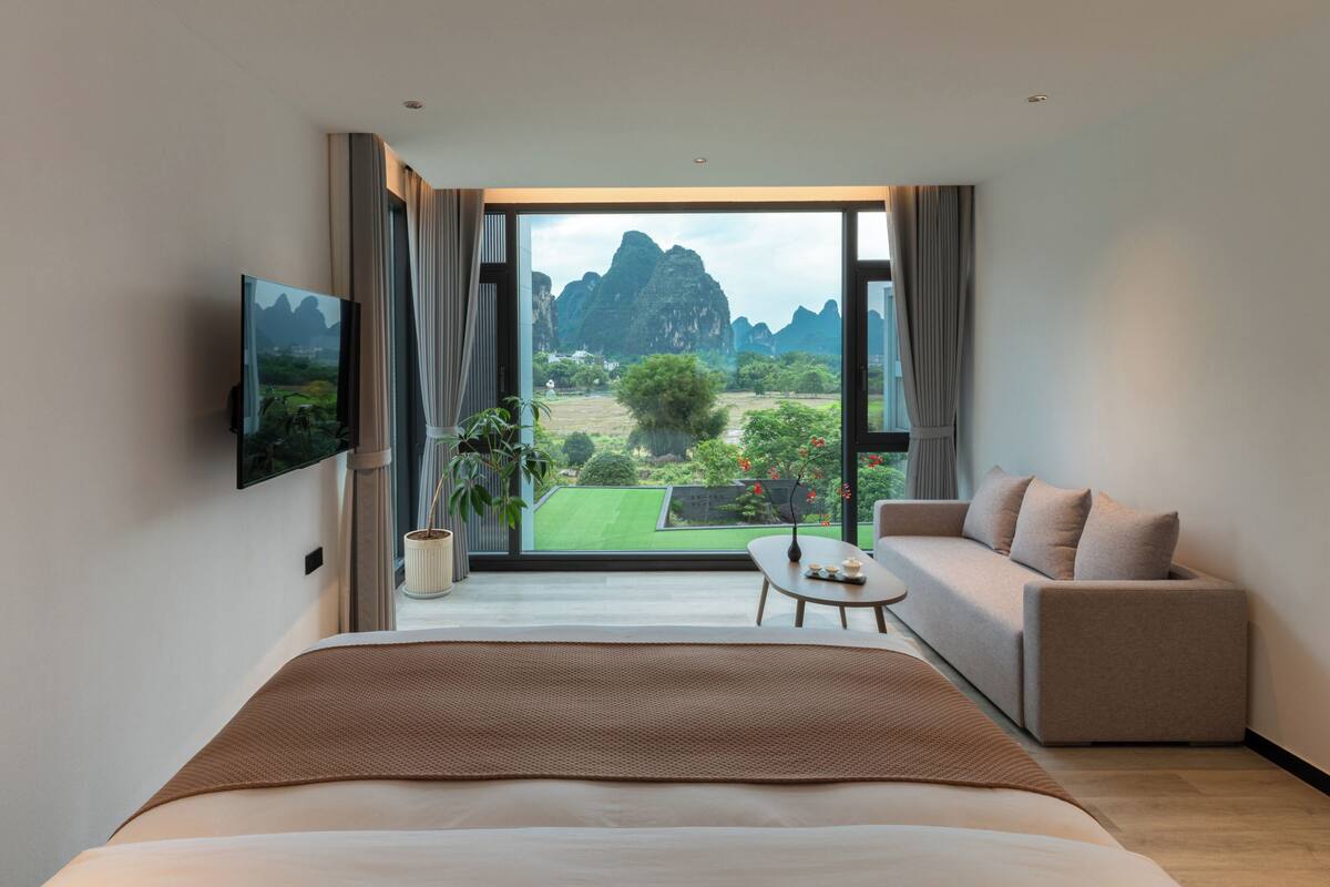 A modern hotel room with a large bed, a sofa, a small table, and a TV. The room has a large window with a view of a mountainous landscape.