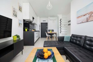 Ett modernt vardagsrum med en svart lädersoffa, ett träbord med frukt på, en platt-TV och ett kök i bakgrunden.