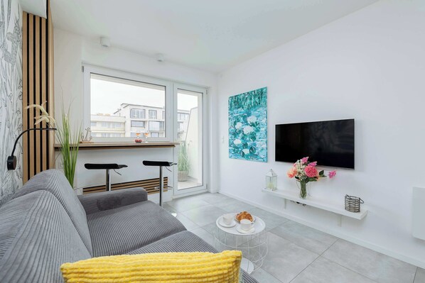 A modern living room with a grey sofa, a yellow throw pillow, a small round coffee table, a TV mounted on the wall, and a window with a view of buildings.