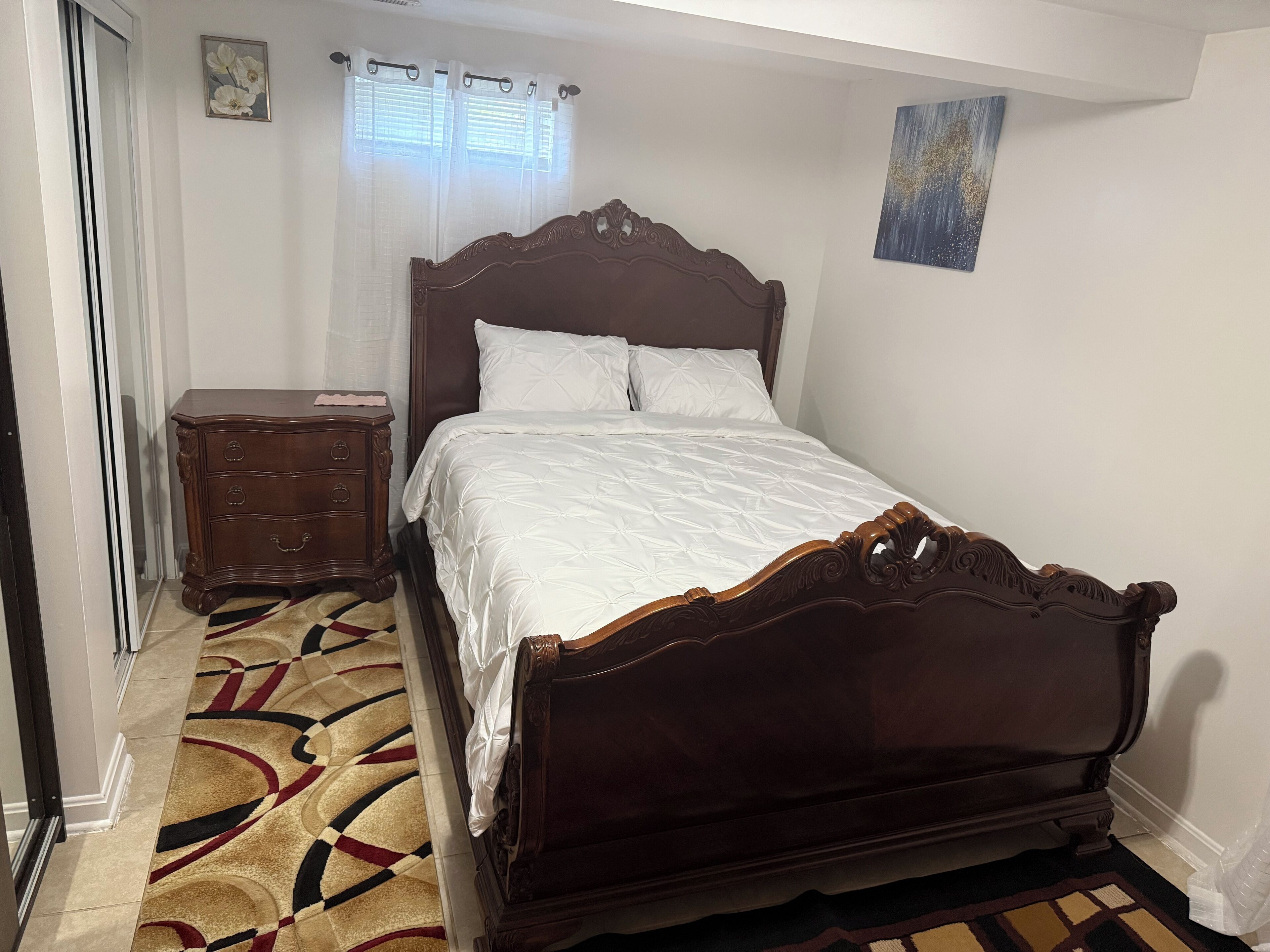A bedroom with a wooden bed, a nightstand, a patterned rug, and a window with sheer curtains.