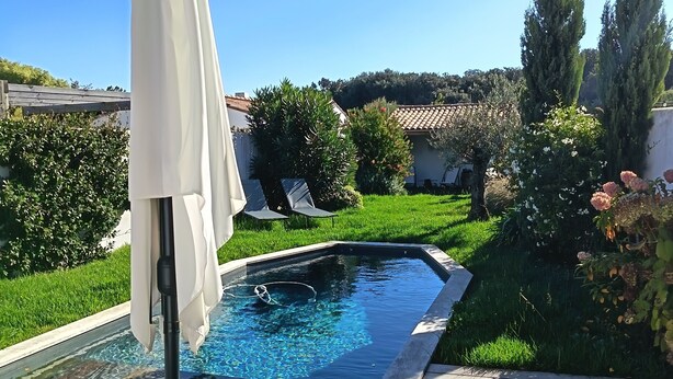 A swimming pool with a wooden deck, surrounded by greenery and a white umbrella. - Villa blanche de Ré (rivedoux)