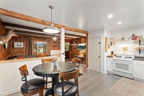 A dining area with a round table and chairs, a wooden ceiling, and a television in the background. - Finger Lakes Cabin | Close to All Top Attractions (Beaver Dams)