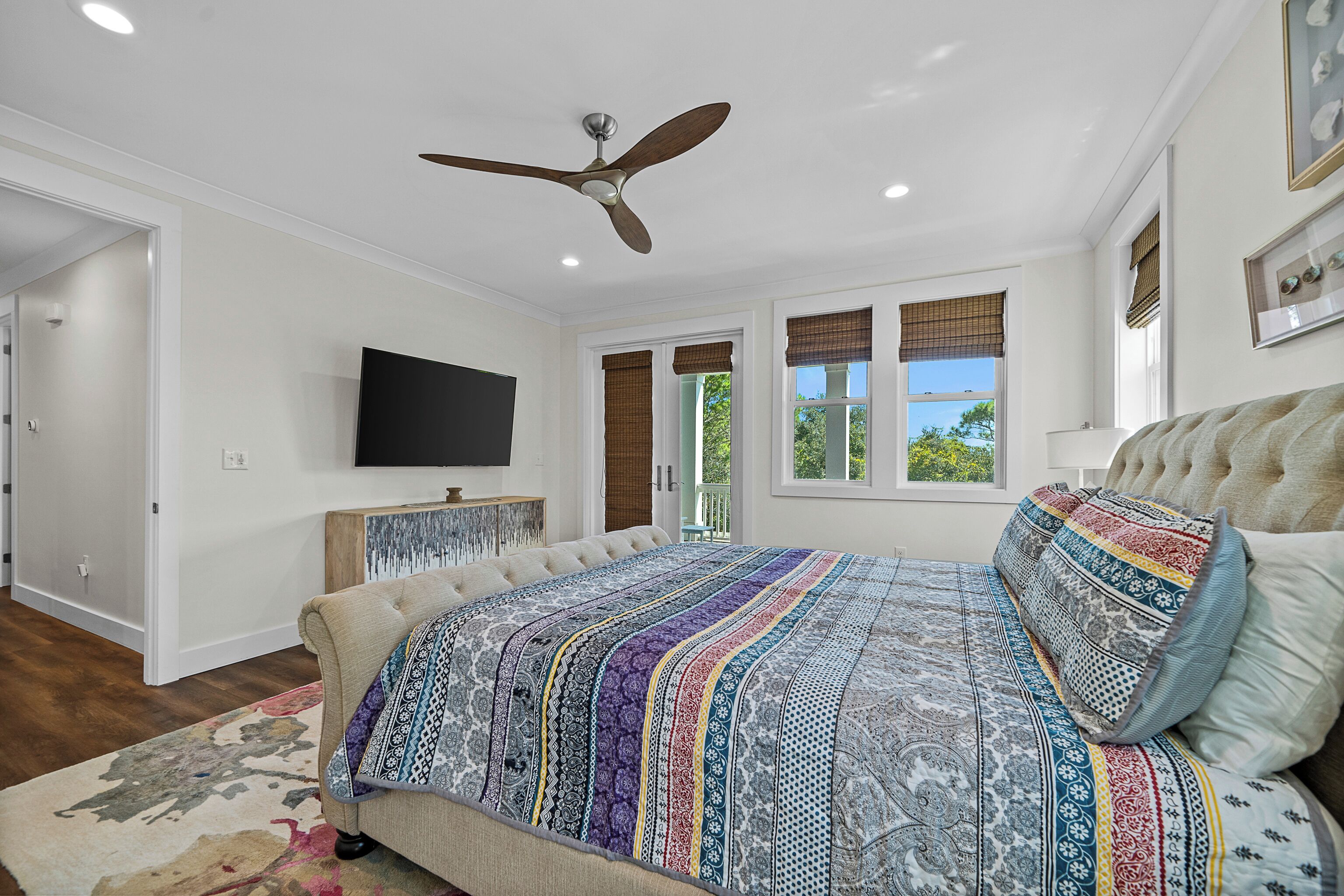 A bedroom with a large bed, a ceiling fan, a flat-screen TV, and a view of the outdoors through a window.
