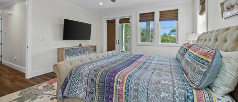 A bedroom with a large bed, a ceiling fan, a flat-screen TV, and a view of the outdoors through a window.