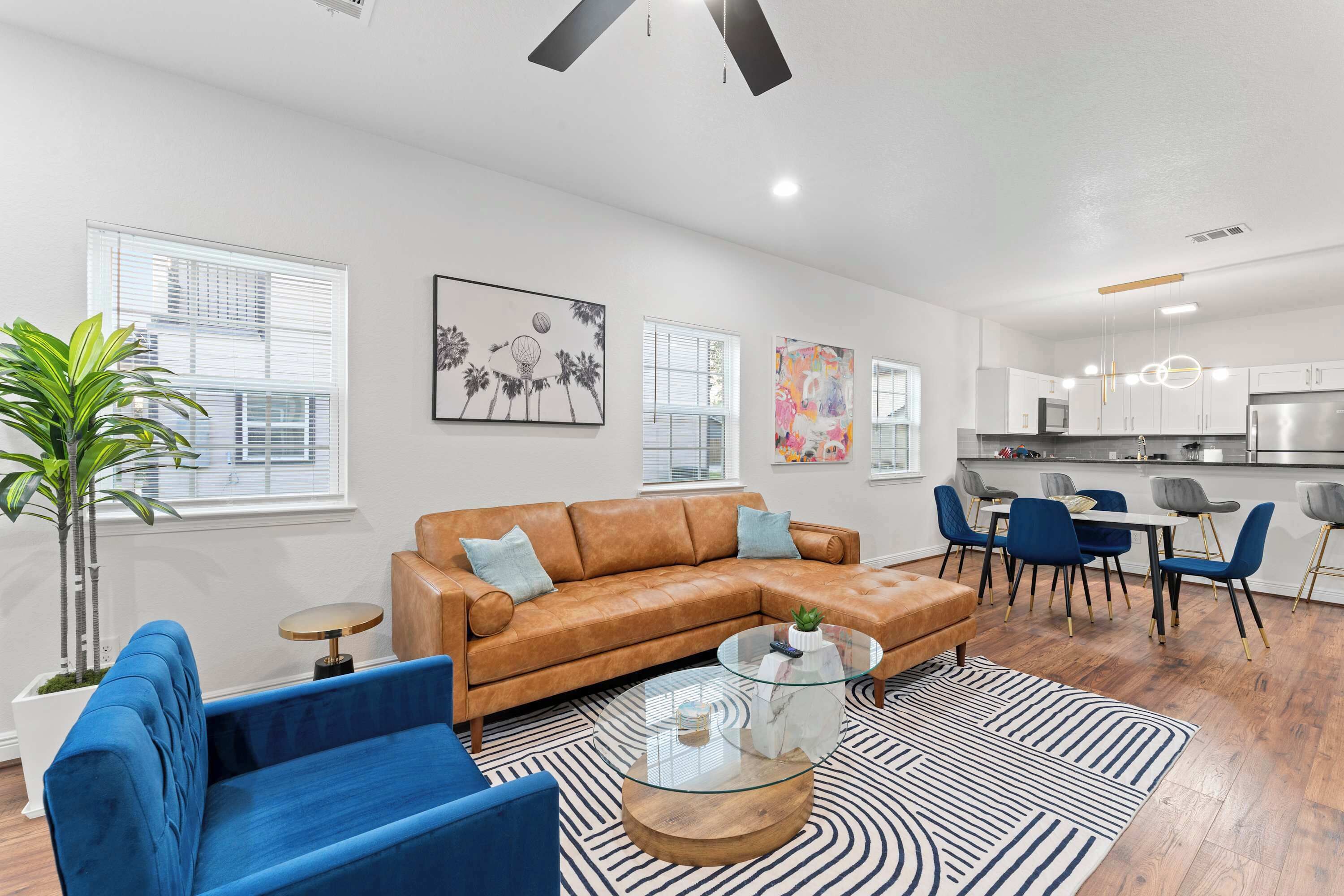 Una sala moderna con un sofá de cuero marrón, un sillón azul y una mesa de centro de vidrio. Hay una zona de comedor con sillas azules y una cocina al fondo.