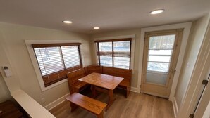 Et spiseområde med et træbord og bænke, store vinduer med persienner, samt en dør med sandblæst glas.