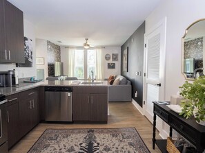 A modern kitchen with dark cabinetry, a dishwasher, and a central island. There is a seating area with a sofa and a dining table. The room has a large window with curtains, a ceiling fan, and a decorative rug.