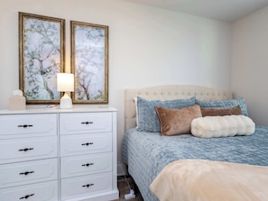 A bedroom with a bed, a bedside table with a lamp, a dresser, and two framed pictures on the wall.