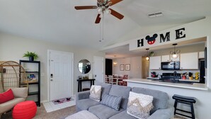 A modern living room with a grey sofa, red ottoman, and a ceiling fan. A kitchen area is visible in the background with a 'HOME' sign above the door. - 5 Mi to Wdw: Resort-style Condo w/ Pool Access (Kissimmee)