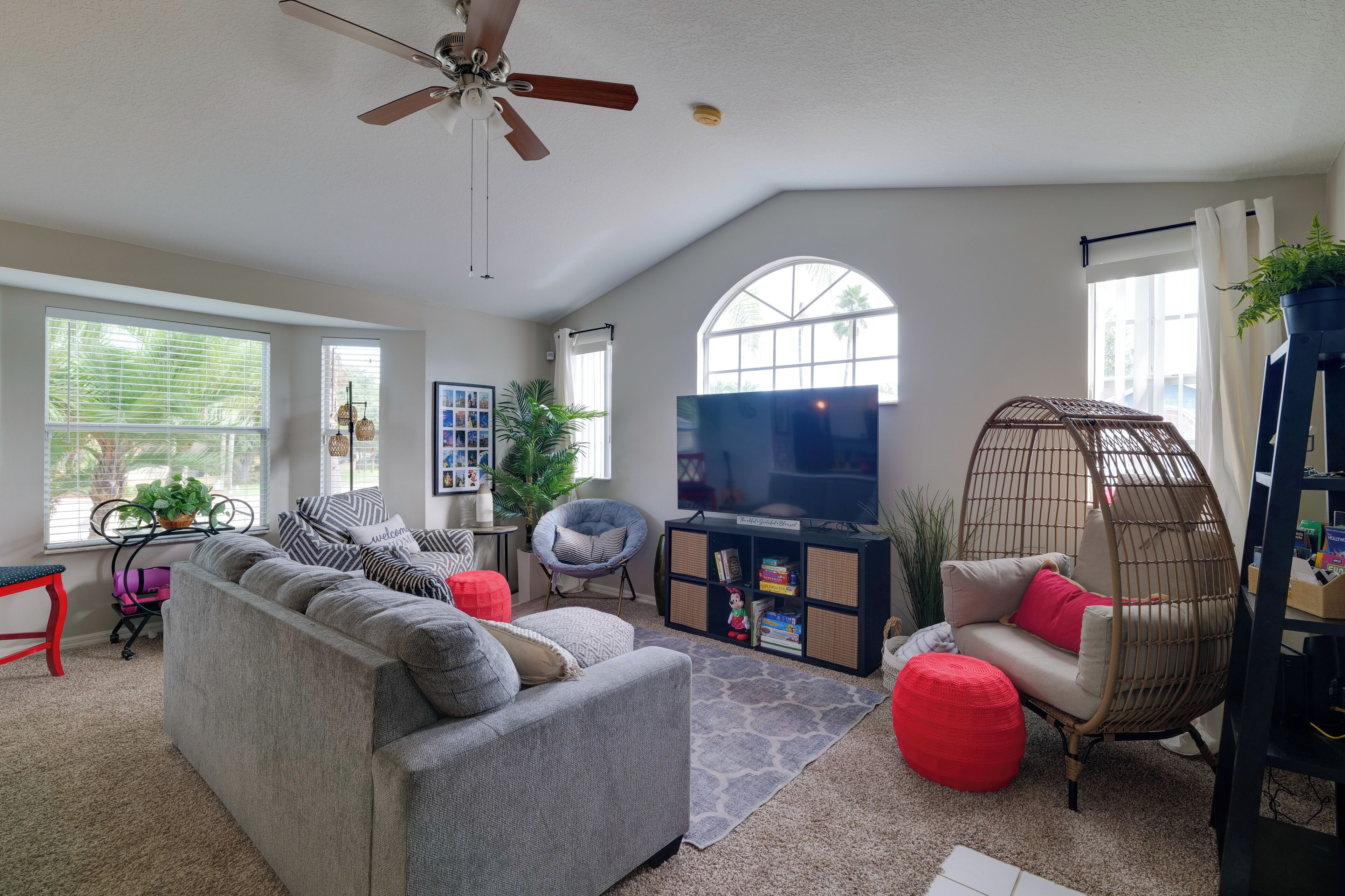 A living room with a grey sofa, red ottoman, wicker chair, and a ceiling fan.