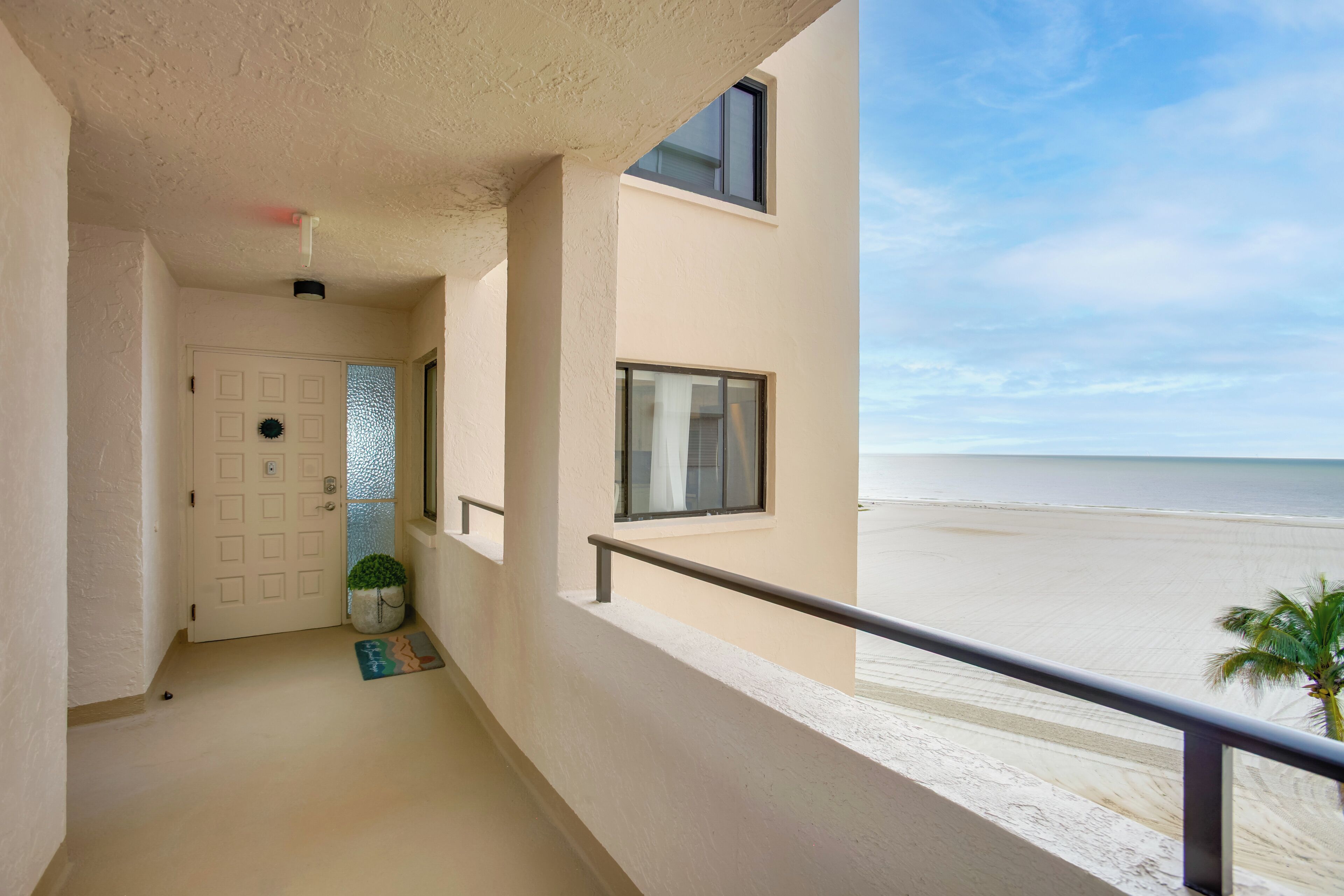 Un balcón con vista a la playa, una puerta con pared texturizada y una planta pequeña en el suelo.