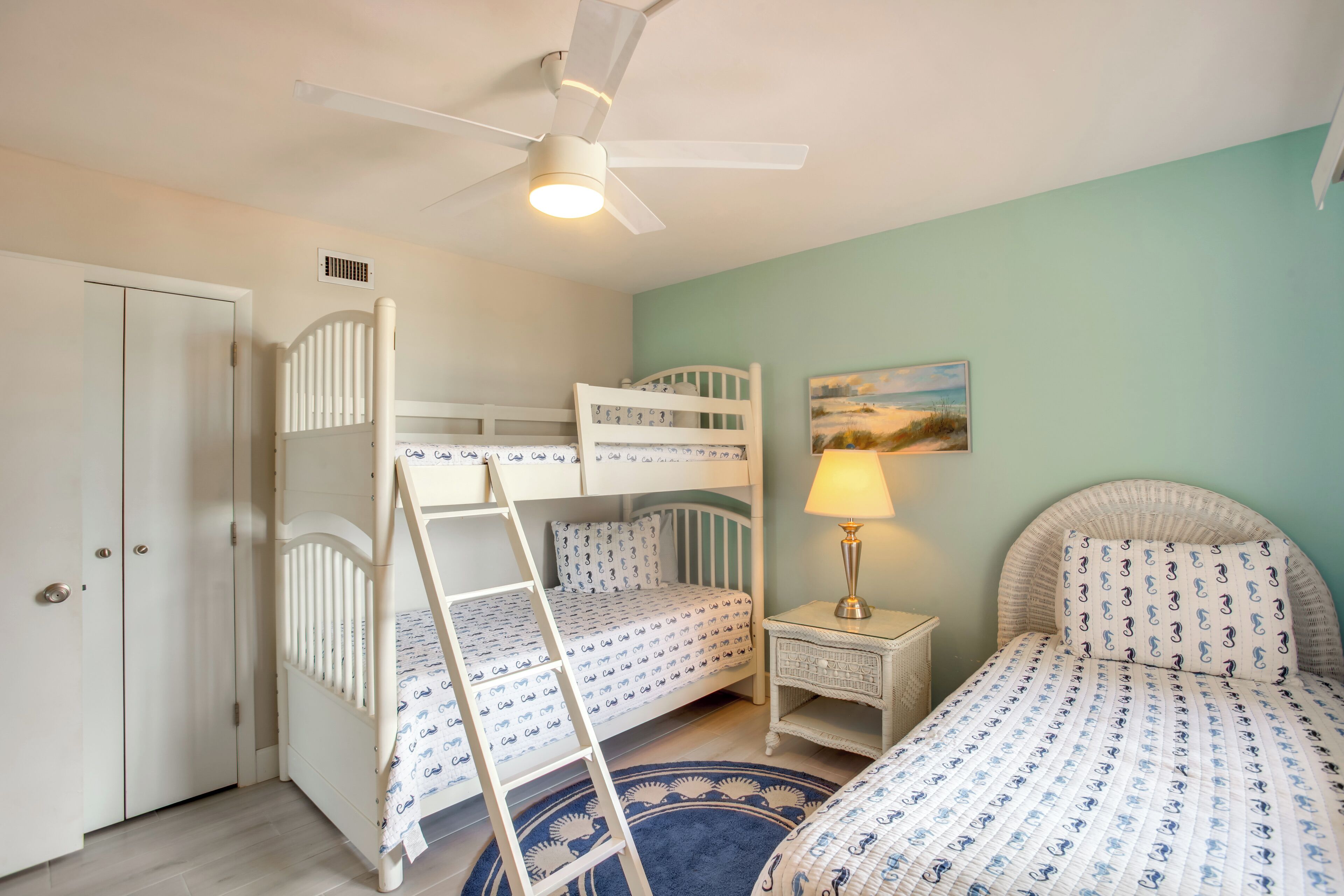 A child's bunk bed room with a ladder, a bedside table with a lamp, and a painting on the wall.