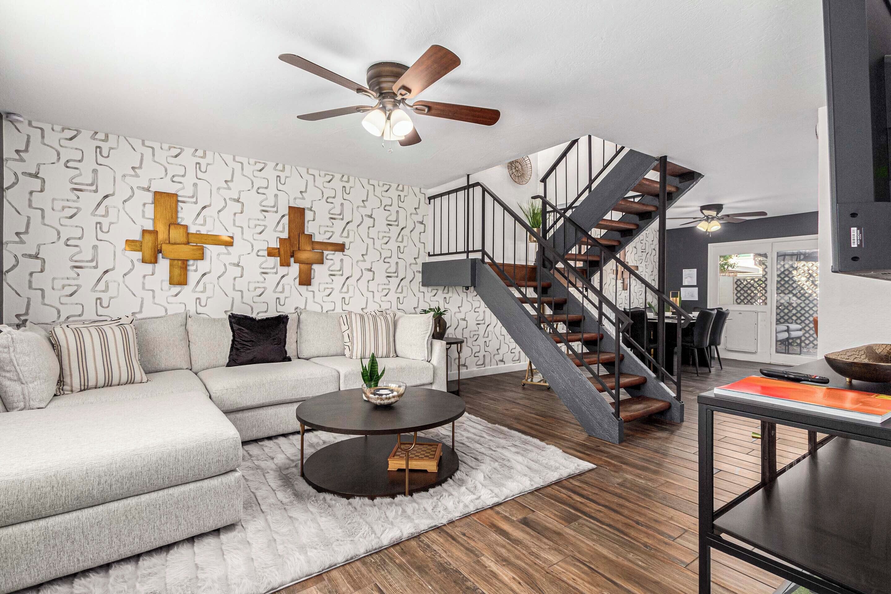 A modern living room with a sectional sofa, a round coffee table, a staircase with a wooden railing, and a ceiling fan.