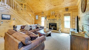 Interior de una cabaña de madera con una escalera, una chimenea, un conjunto de sofás y un televisor.