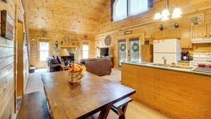 Interior de una cabaña de madera con mesa de comedor, sofá, zona de cocina y chimenea.