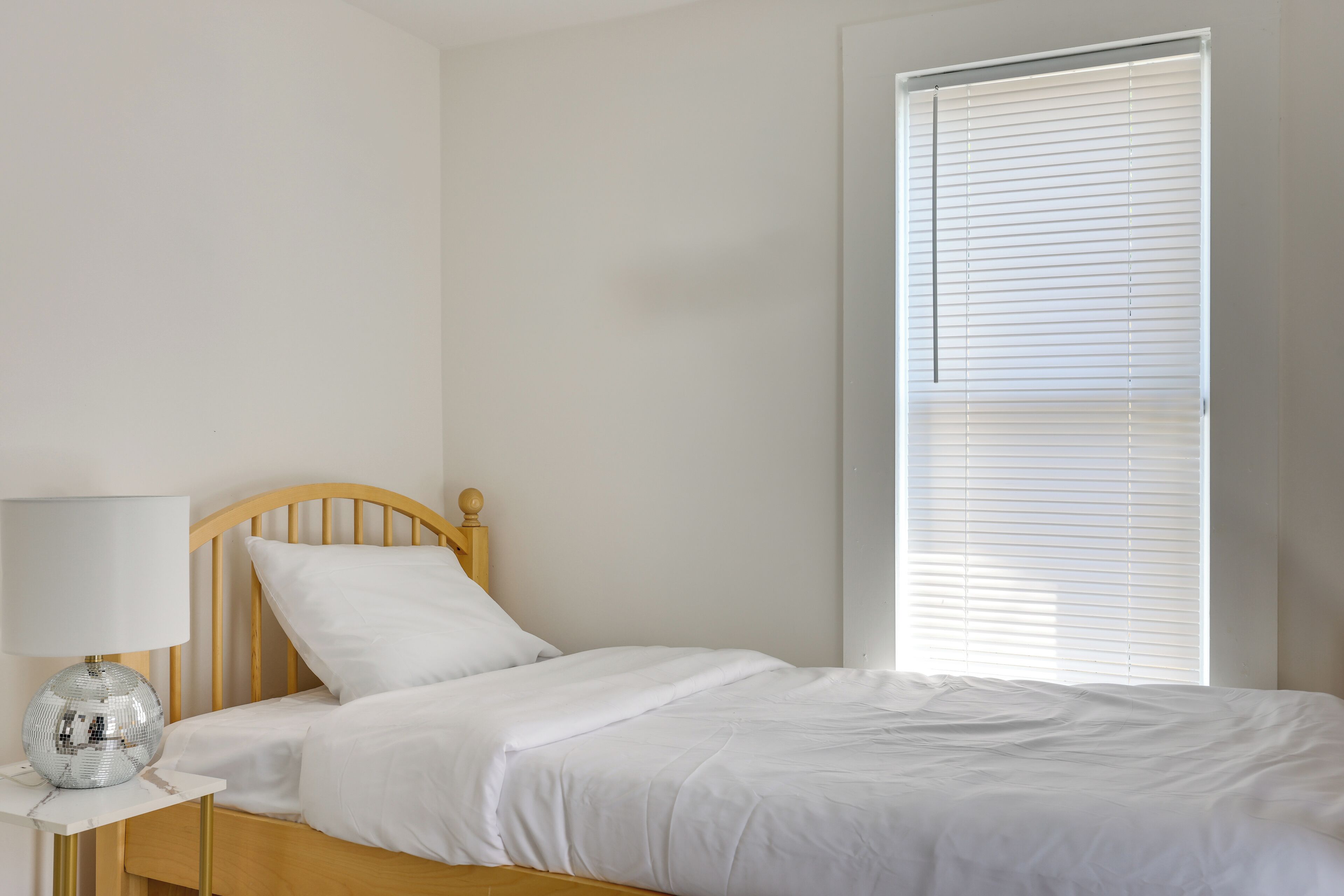 A bedroom with a wooden bed, white bedding, a bedside table with a lamp, and a window with blinds.