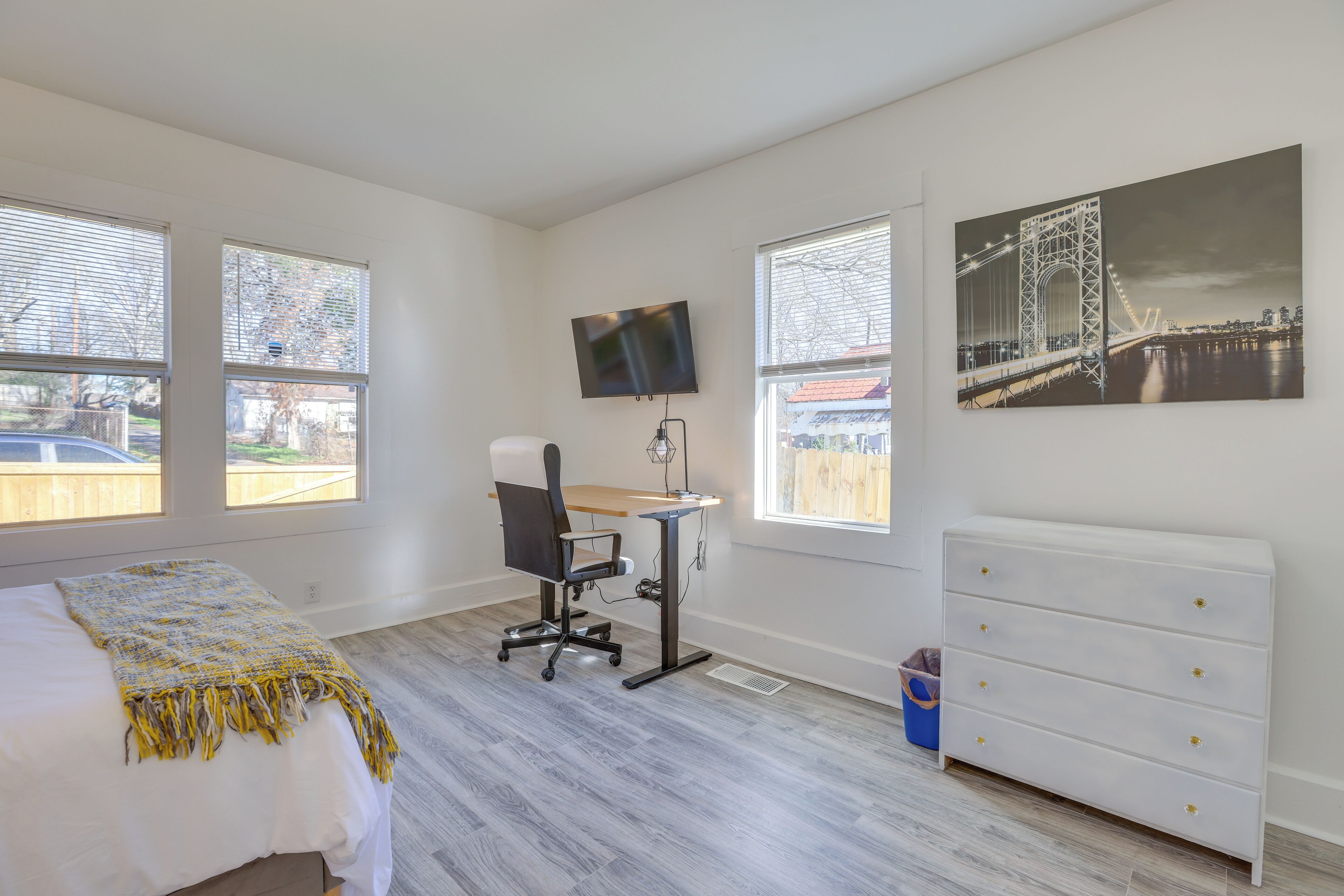 Un dormitorio con cama, escritorio, silla, aparador, televisor y ventanas amplias.