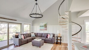 Amplia sala de estar con sofá modular, una mesa de centro y un gran ventanal con vistas al exterior. Se observa una escalera de caracol al fondo.