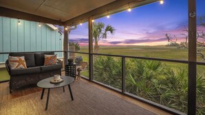 Un balcón con sofá y mesa, ventanas amplias con luces de cuerda y vistas a un campo y al atardecer.