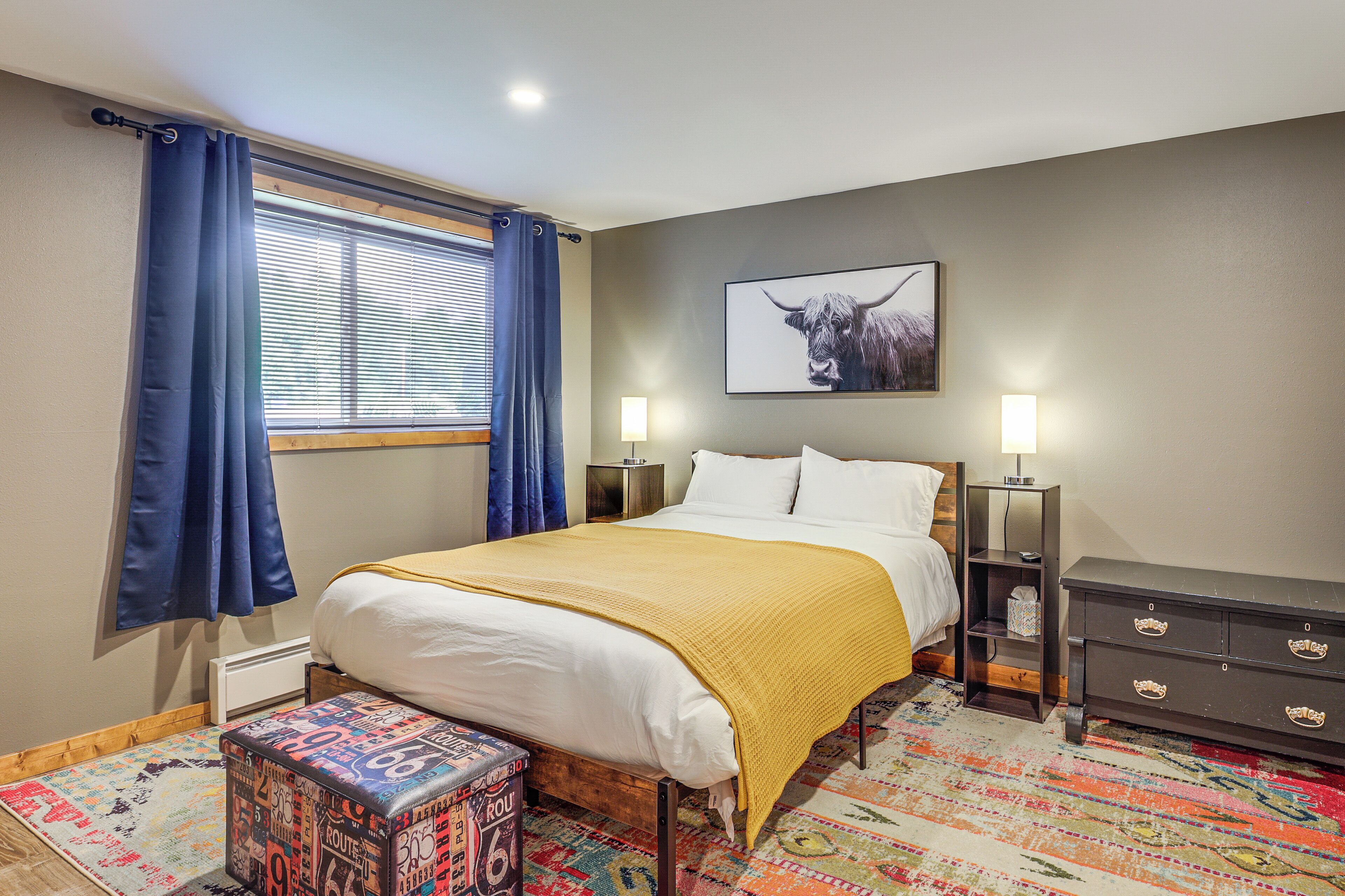 Une chambre à coucher comprenant un lit, une table de chevet, une commode, une lampe, un tapis et une fenêtre avec des rideaux.
