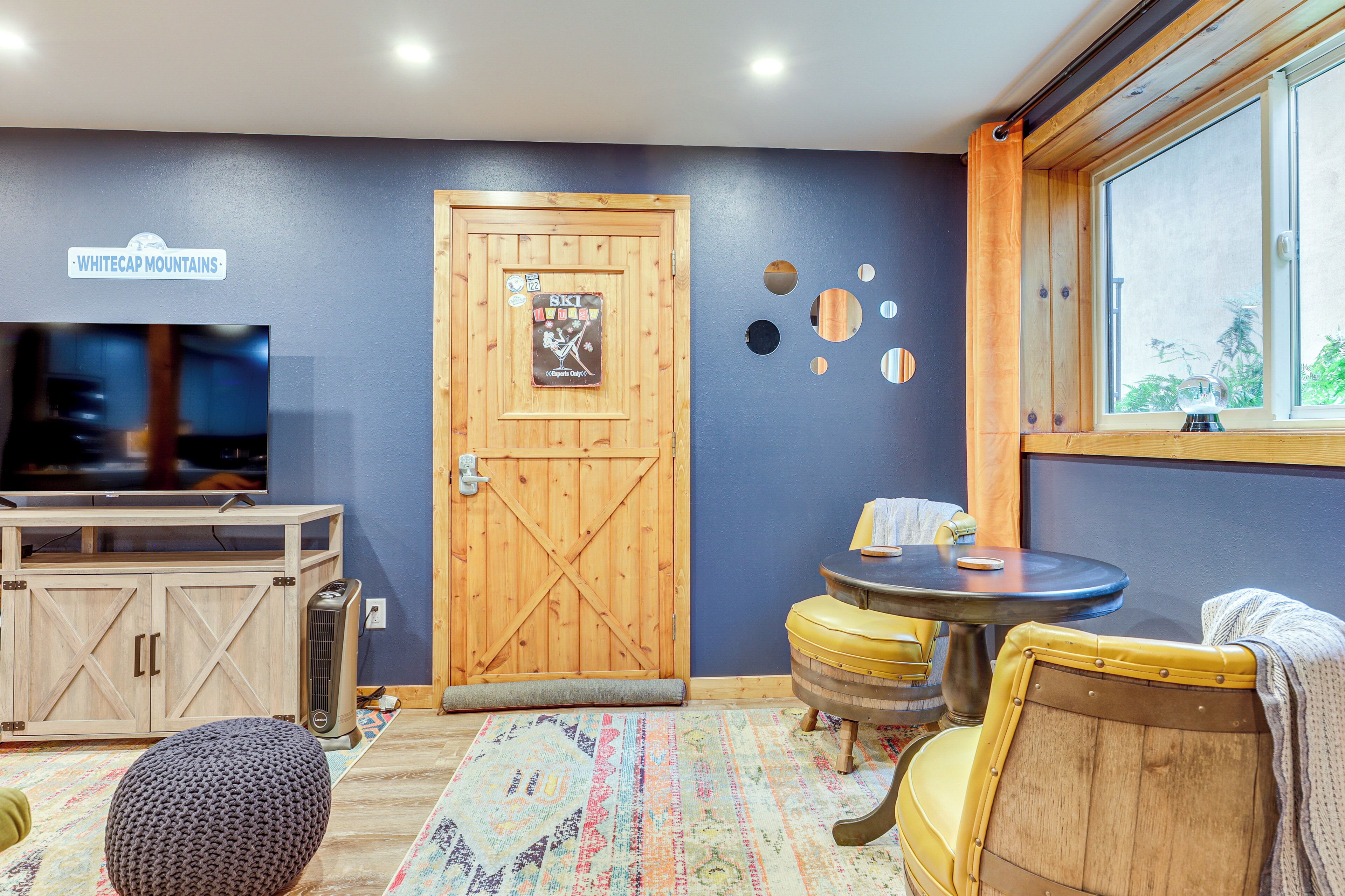 A room with a wooden door, a television, a small table, a yellow chair, and a patterned rug.