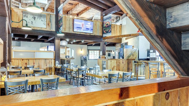 Restaurante com interiores de madeira, várias mesas e cadeiras para refeições, e uma televisão exibindo um anúncio.