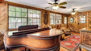A wooden interior with a piano, a ceiling fan, and a large window with blinds. - Sandy Shore & Multi-boat Dock! St Johns River Home (Astor)