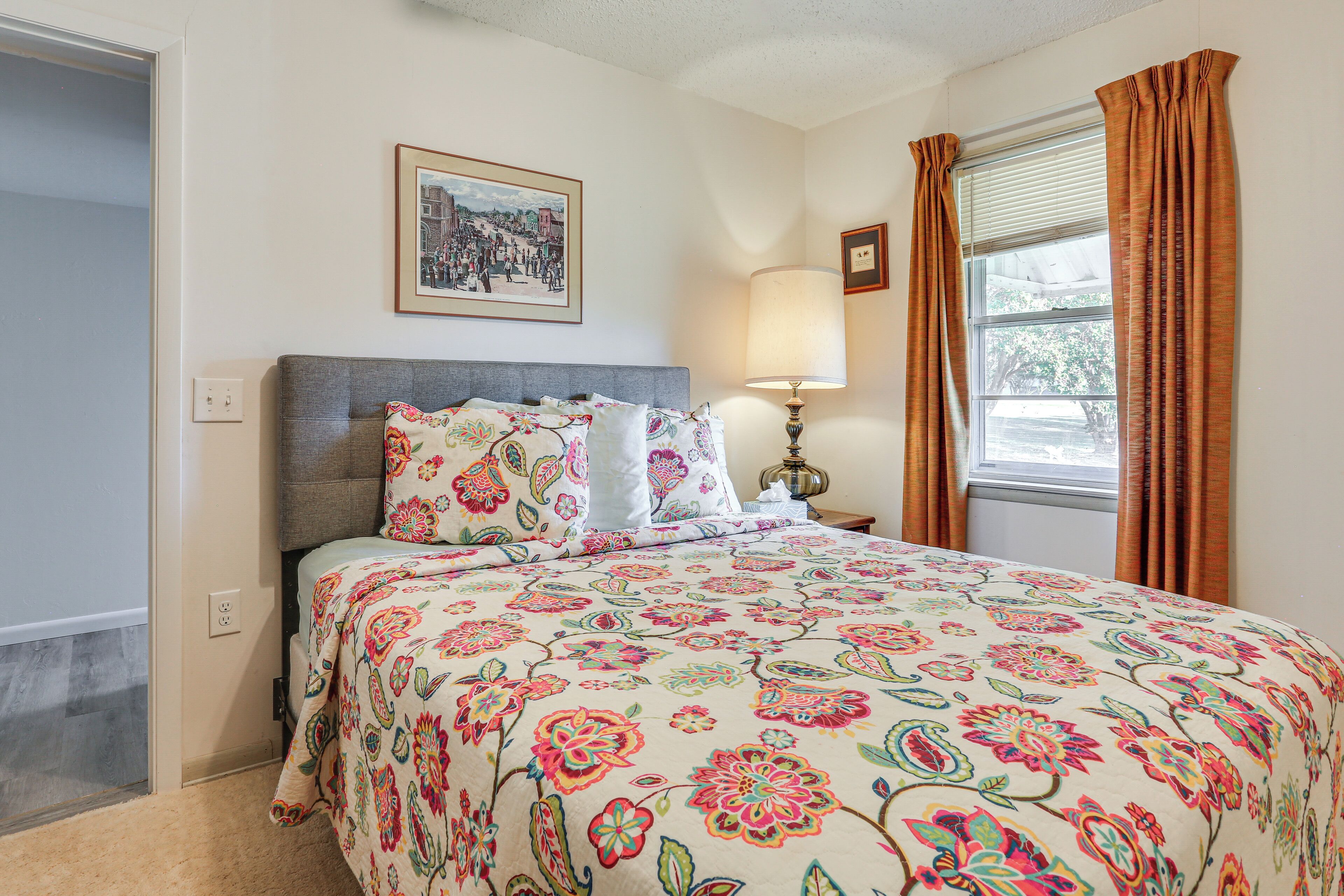 Un dormitorio con cubrecama floral, una lámpara de noche, un cuadro enmarcado en la pared y una ventana con cortinas.