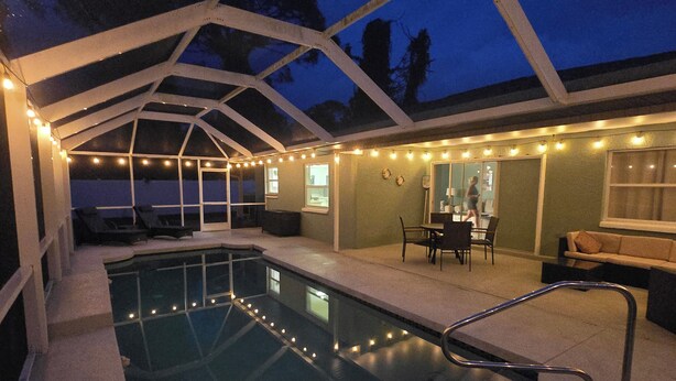 A covered outdoor swimming pool area with steps leading into the pool, a seating area with chairs and a table, and string lights along the perimeter. - 3 Mi to Manasota Key: Stylish Single-story Home! (Venice)