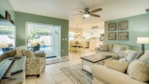 A spacious living room with a sofa set, a coffee table, and a dining area with chairs. There is a ceiling fan, a kitchen in the background, and a door leading outside. - 3 Mi to Manasota Key: Stylish Single-story Home! (Venice)