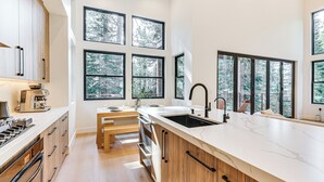 A modern kitchen with a large island, built-in oven, and a view of the forest through large windows. - 4 Mi to Soda Springs Resort: Hot Tub Hideaway! (Truckee)