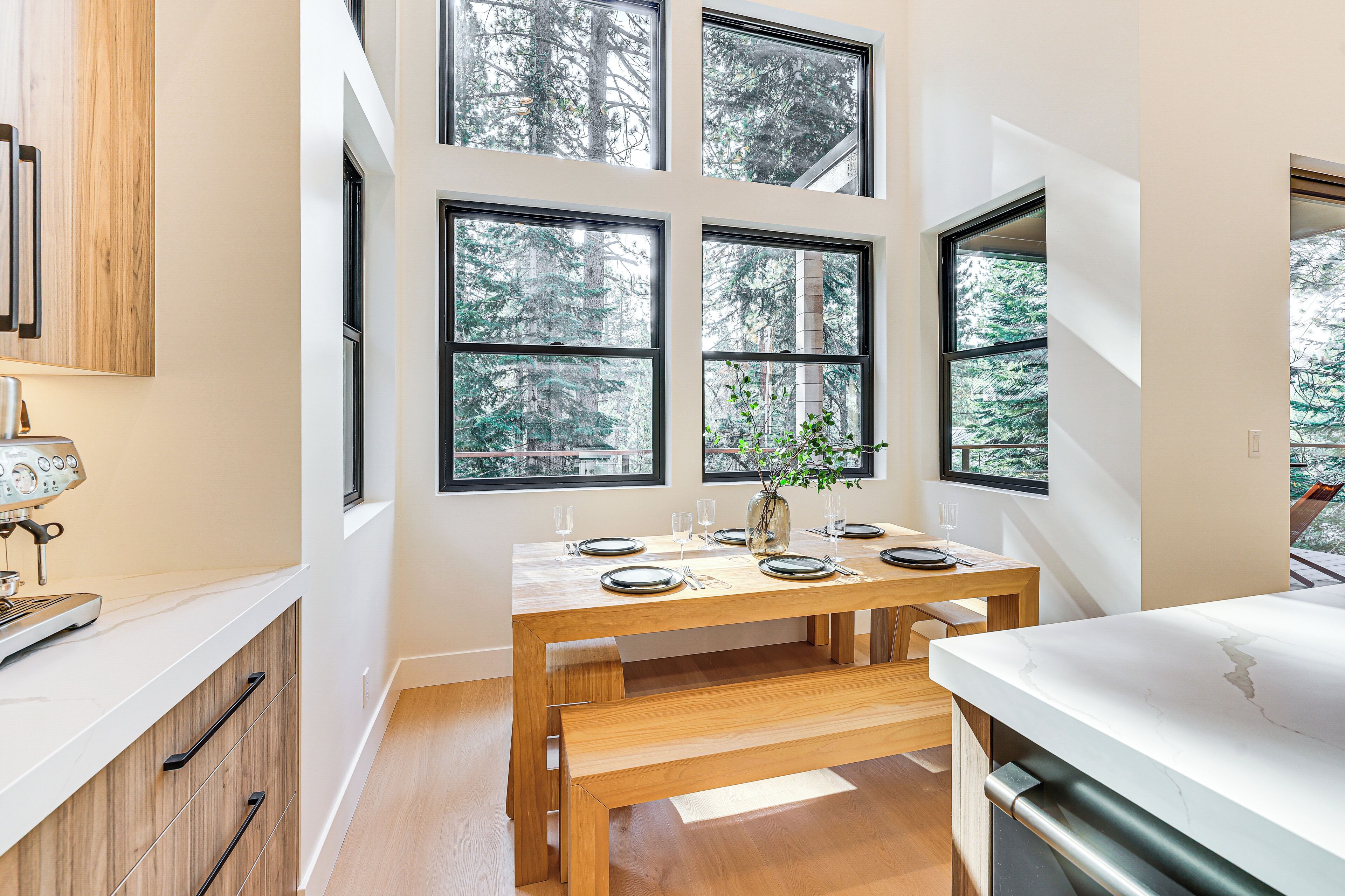 Eine moderne Küche mit Holztisch, gedeckt für vier Personen, großen Fenstern mit Blick auf einen Wald und einer integrierten Espressomaschine.