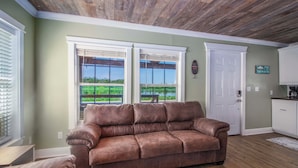 A living room with a brown leather sofa, a wooden ceiling, and a view of a green landscape through large windows. - Private Dock: Des Allemands Cabin on the Bayou! (Des Allemands)
