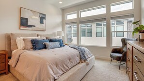 A bedroom with a large bed, a nightstand, a dresser, a chair, and a window with blinds.