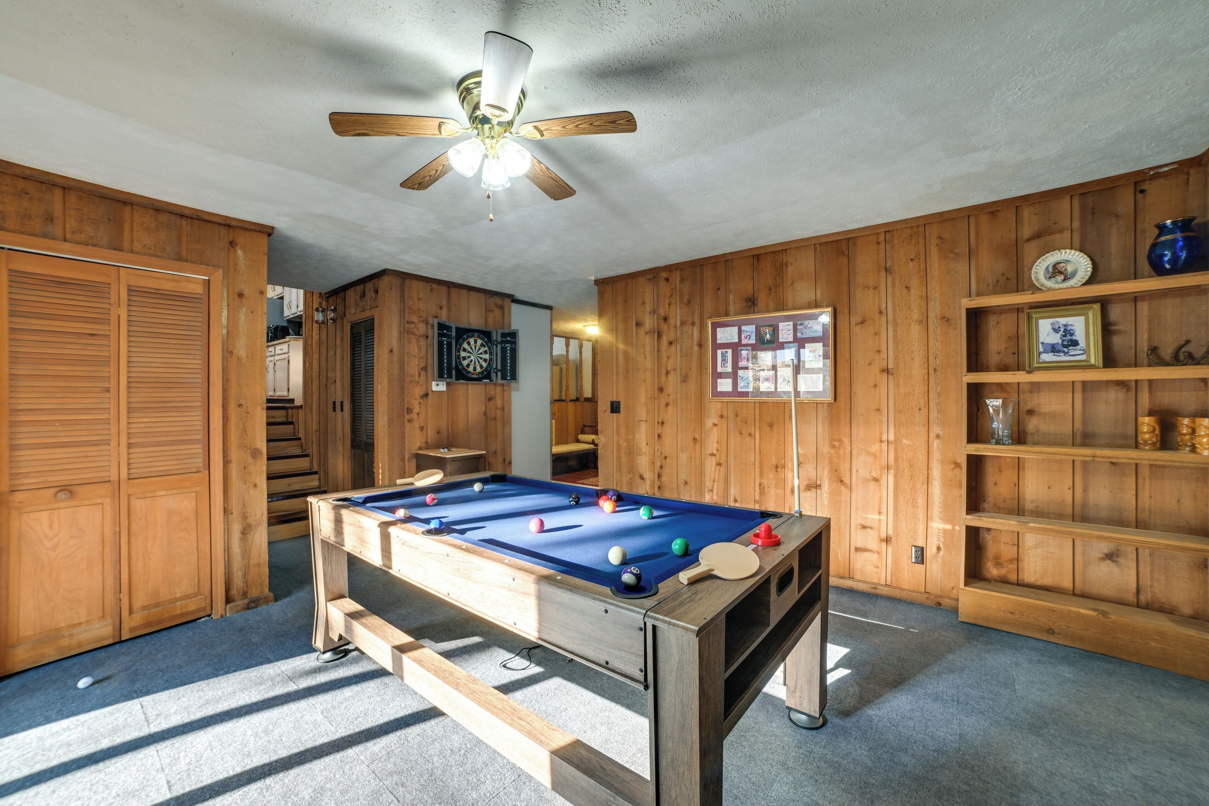 Une table de billard dans une pièce aux murs en bois, avec un ventilateur au plafond et des étagères remplies de divers objets.