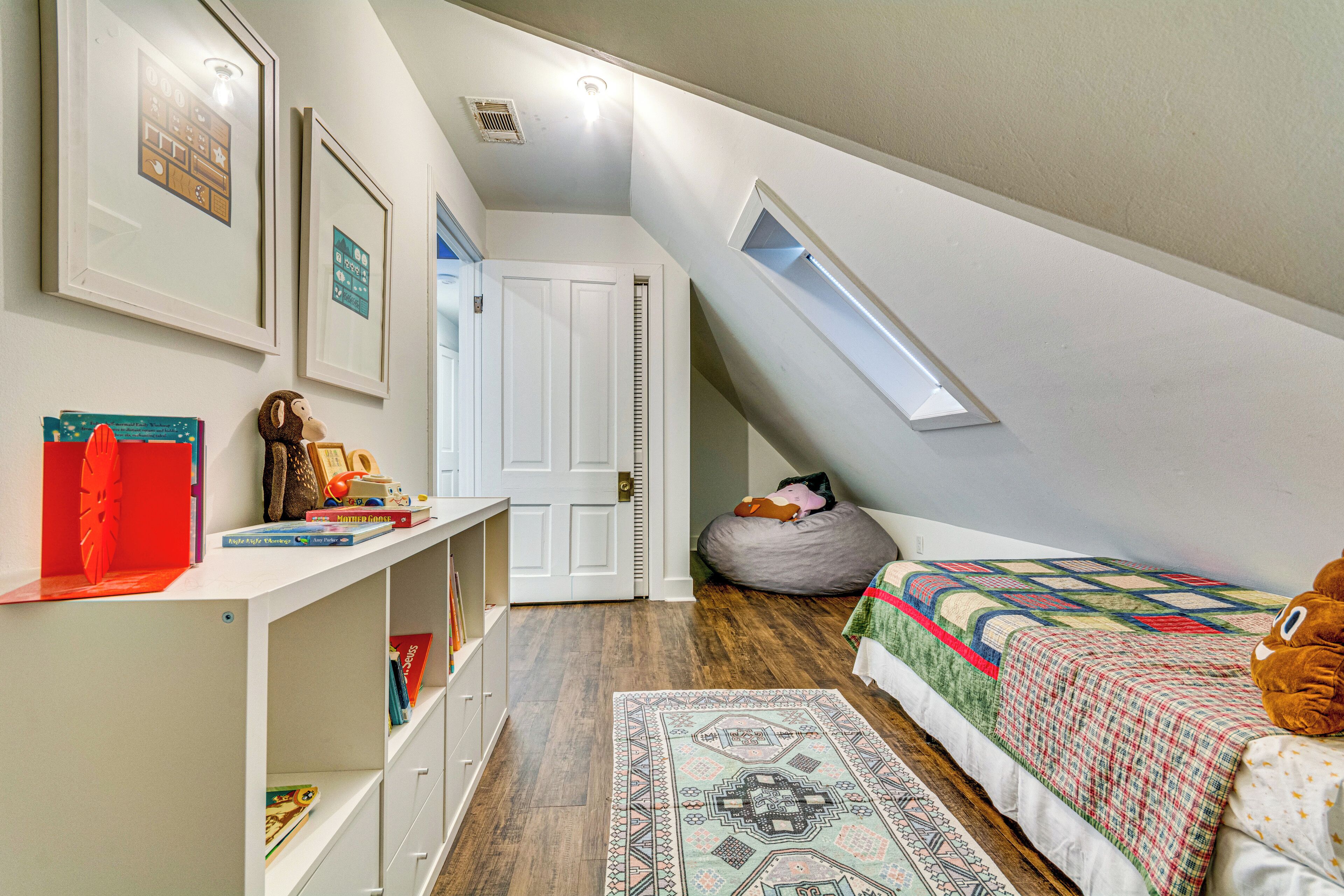 Un dormitorio con techo inclinado, tragaluz, cama con colcha, sillón de mimbre, estantería y una puerta.