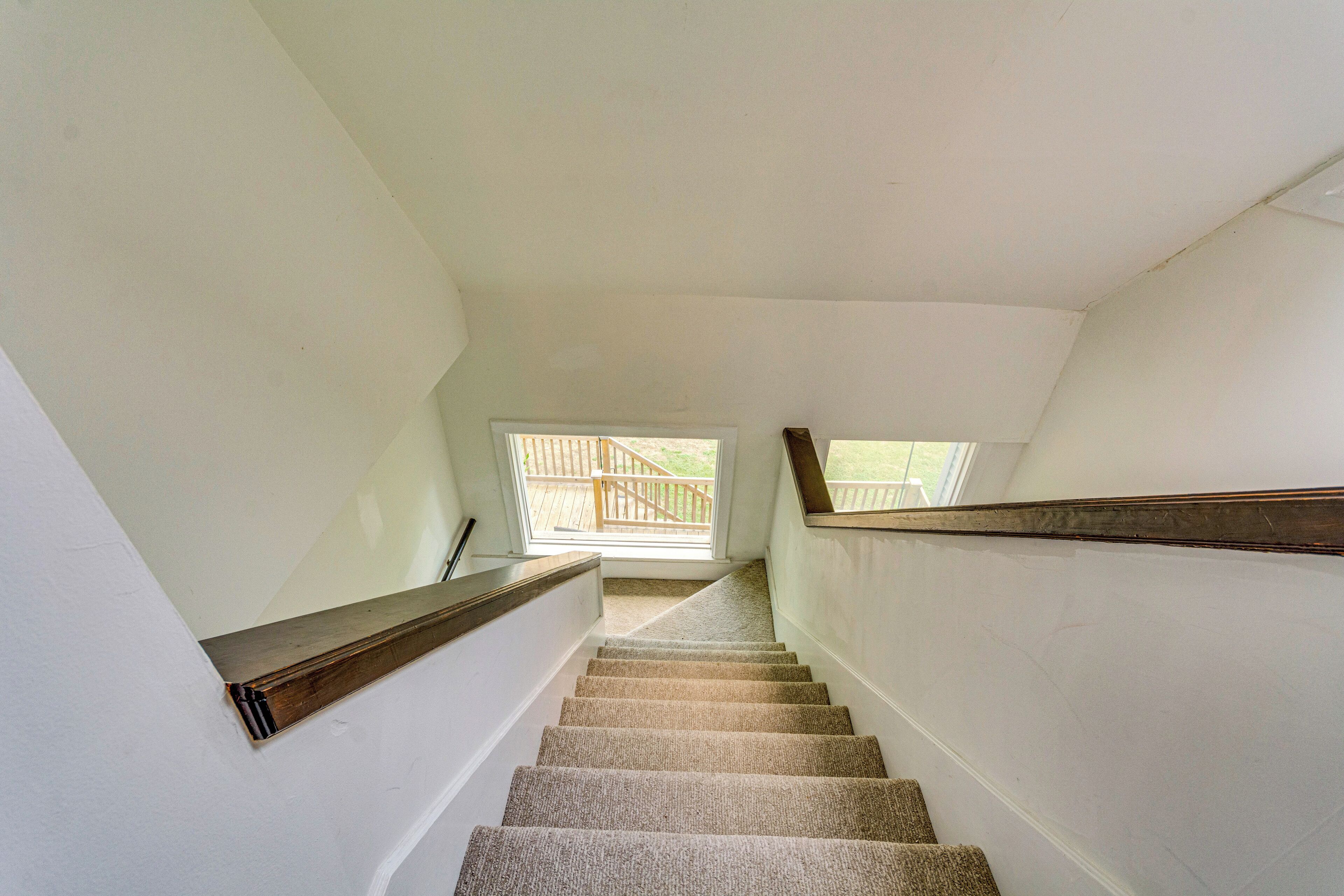 A carpeted staircase with wooden handrails, leading to a bright room with a window and a view of a deck outside.