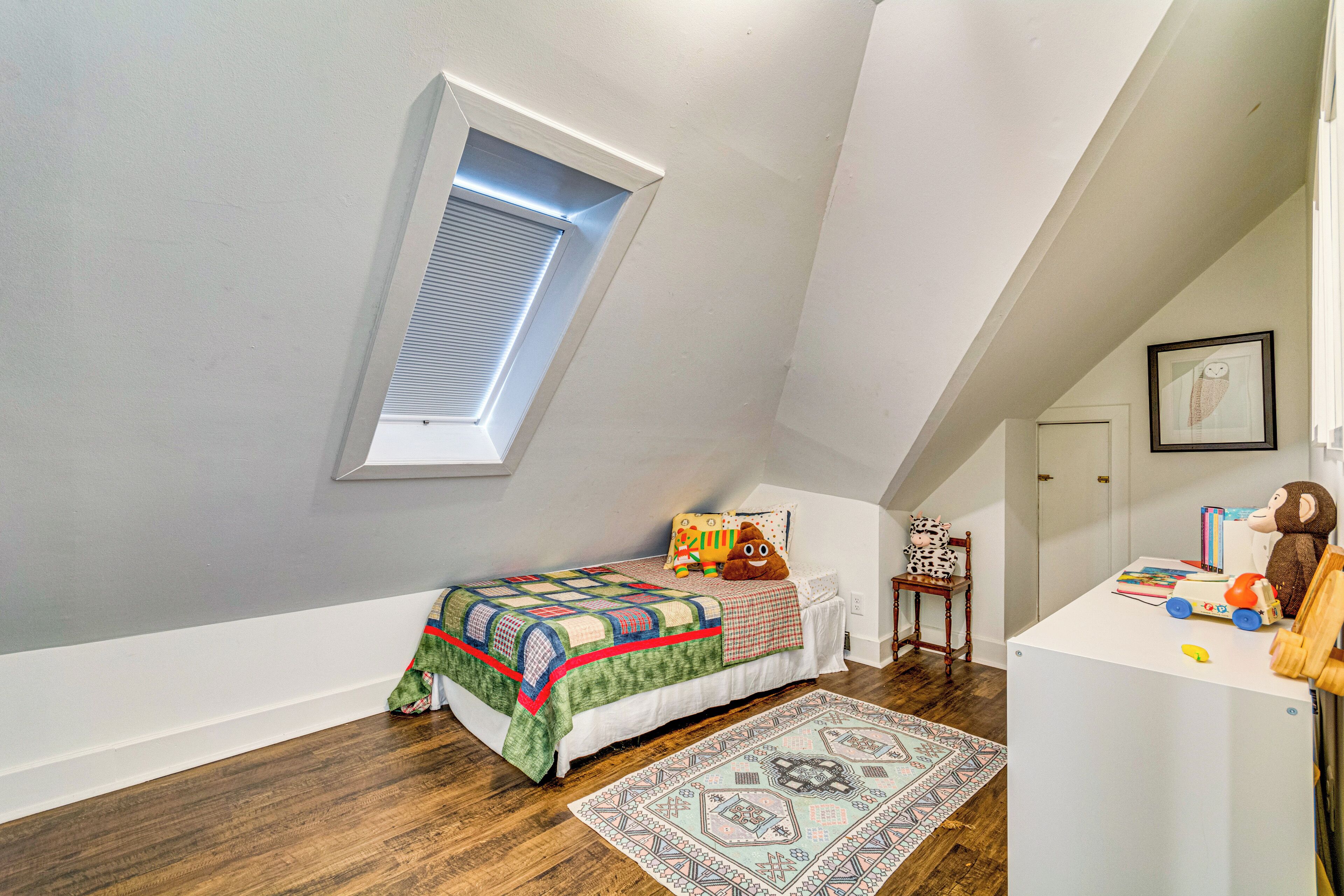 A bedroom with a sloped ceiling, a skylight, a bed with a colorful quilt, a small desk with toys, and a patterned rug.