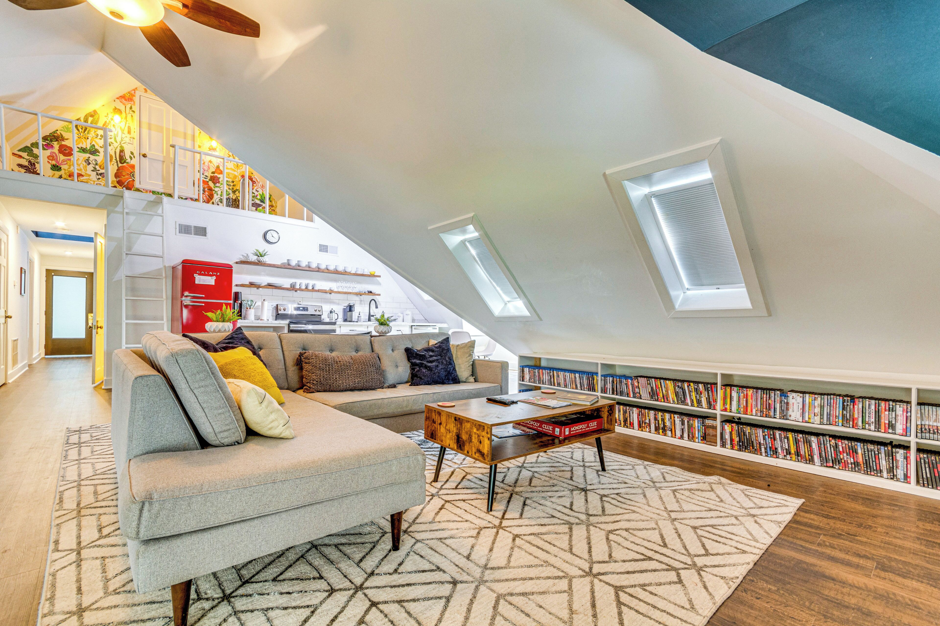 A spacious loft apartment with a high ceiling, a built-in bookshelf, a cozy seating area with a sofa and coffee table, and a kitchen in the background.