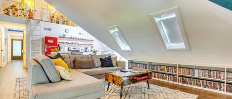 A spacious loft apartment with a high ceiling, a built-in bookshelf, a cozy seating area with a sofa and coffee table, and a kitchen in the background.