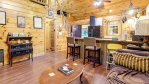 Ein gemütliches Blockhaus-Interieur mit Holzausstattung, einer Essecke mit zwei Stühlen, einem Tisch mit einem Buch und einer Küche mit Kühlschrank und Herd.
