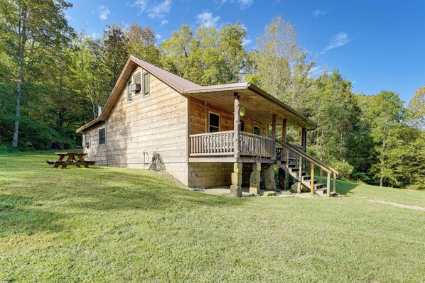 A wooden cabin with a porch and a picnic table in a grassy area surrounded by trees.