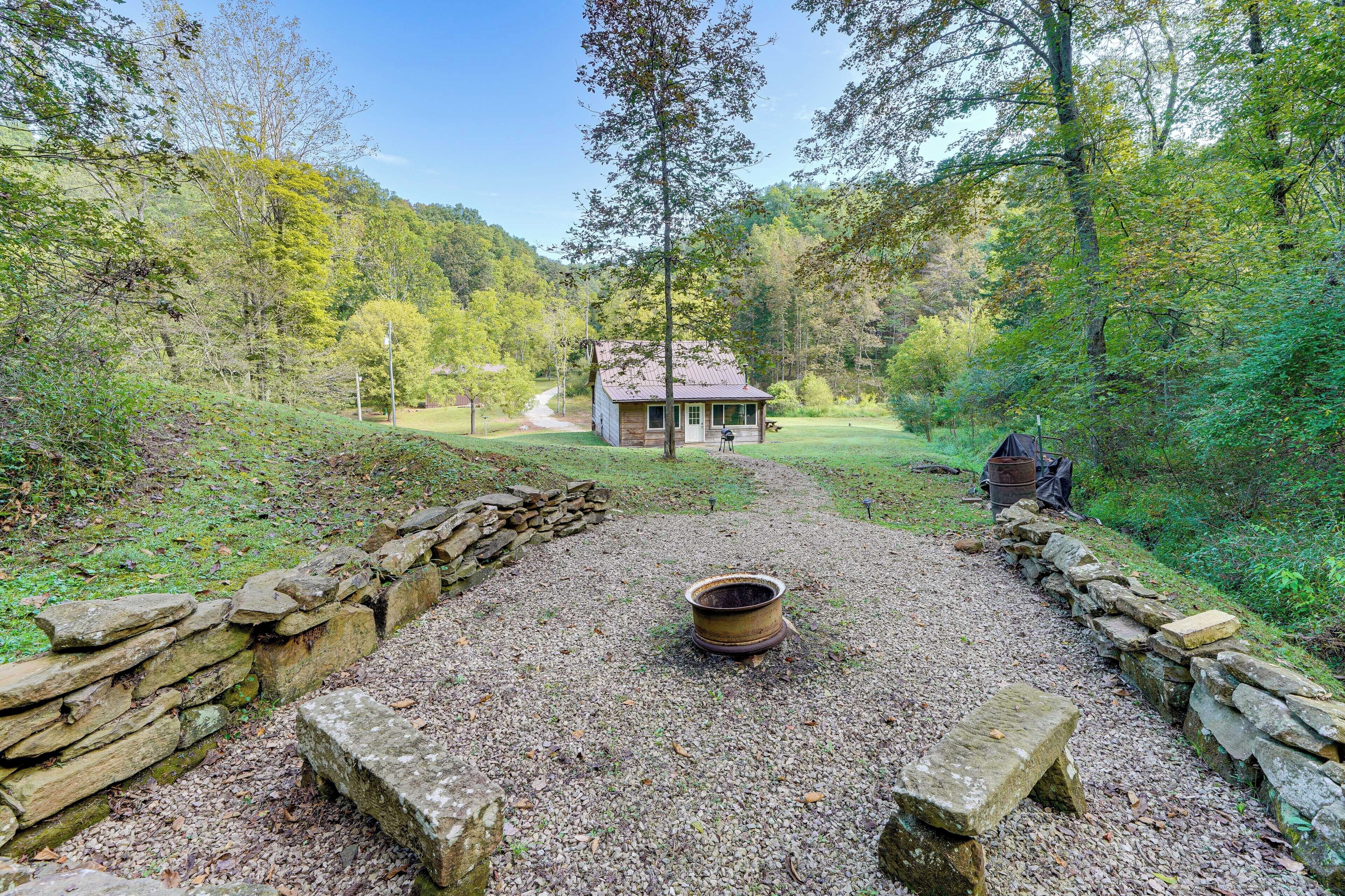 A rustic cabin with a stone wall and a fire pit in the center, surrounded by a gravel area and dense forest.
