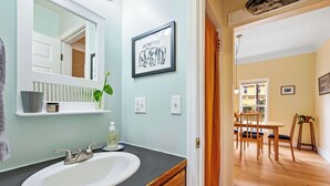 Une salle de bain avec un lavabo blanc, un miroir, un tableau encadré et une plante posée sur le comptoir.