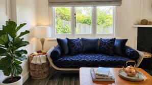 A cozy living room with a dark blue patterned sofa, a potted plant, a small table with a book and a teapot, and a window with a view of greenery. - Bowen Toft Villa- home-from-home (FIFO friendly) (Sandy Bay)