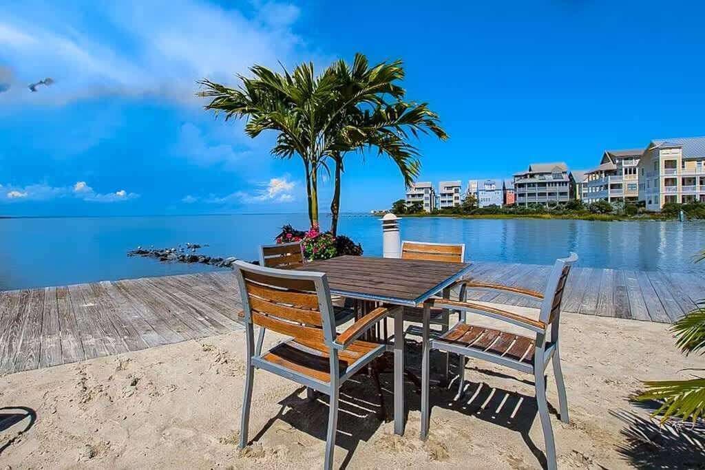 A wooden deck with a table and chairs, palm trees, and a view of the ocean and buildings in the background.