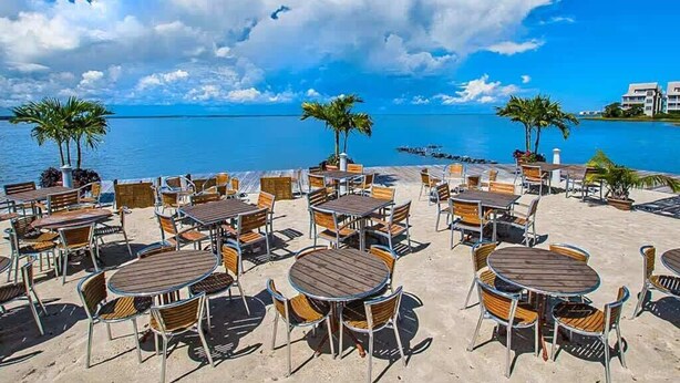 An outdoor seating area with round tables and chairs, palm trees, and a view of the ocean. - 5 Corner Store Ln Sunset Island | Resort Community (Ocean City)