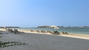 A beach with rows of sun loungers and umbrellas, a clear sky, and a distant view of buildings. - Relaxing Beach Access 1 Bedroom in Grand Bleu (Dubai)