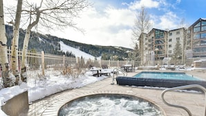 Ett bubbelbad omgivet av en snötäckt terrass, en pool och en bergig bakgrund.