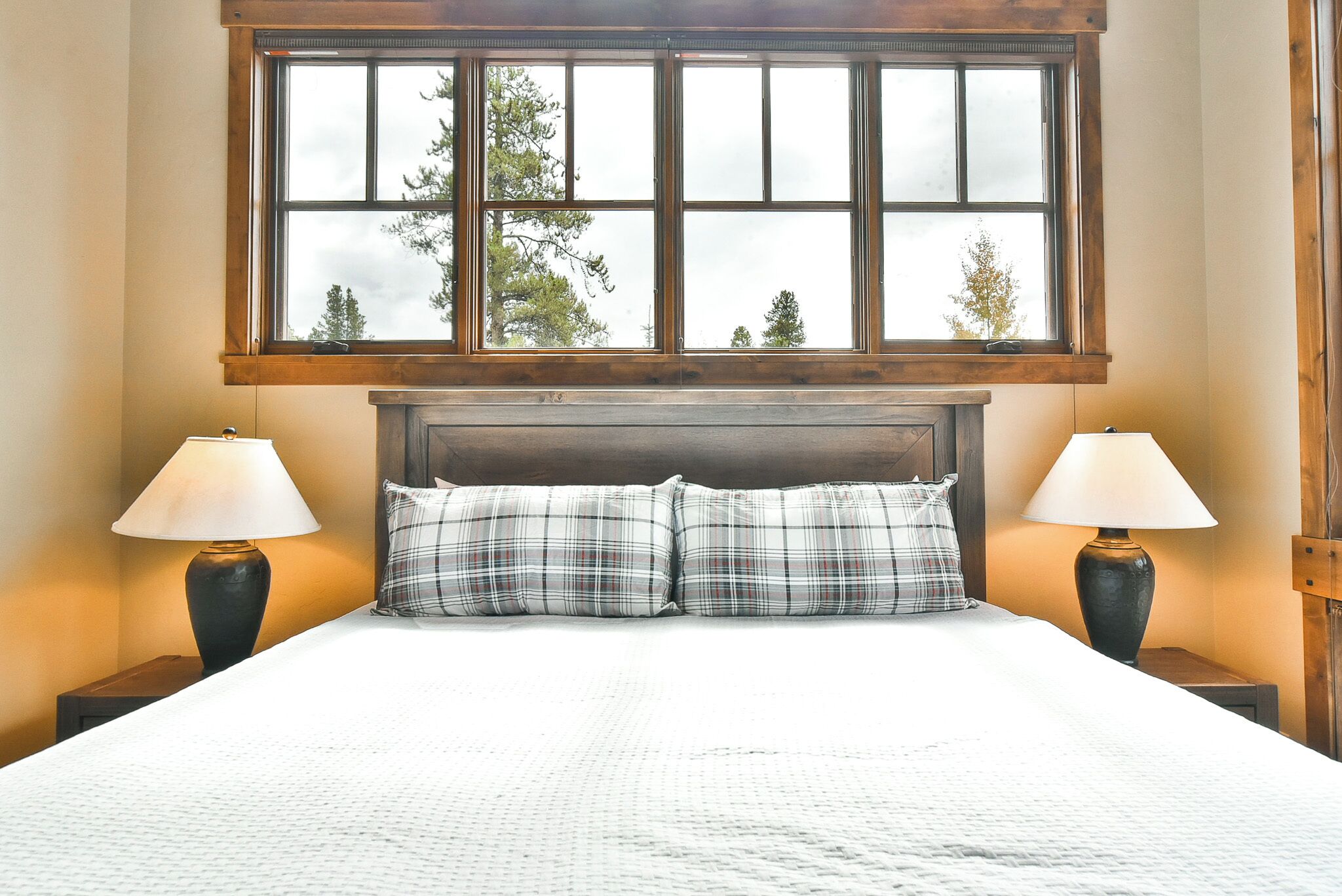A neatly made bed with plaid pillows, two bedside lamps, and a large window with a view of trees.